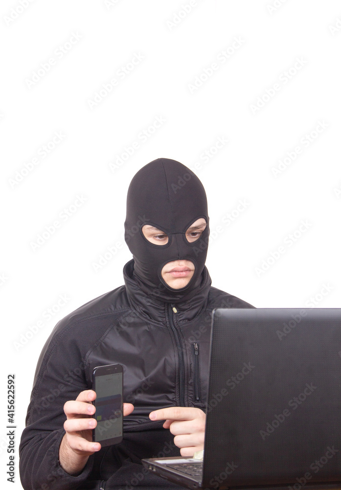man in black mask with phone and laptop engaged in internet fraud man in black mask with phone and laptop engaged in internet fraud