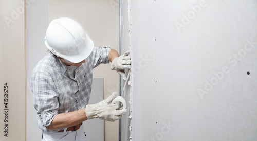 man drywall worker or plasterer putting mesh tape for plasterboard on a wall using a spatula and plaster. Wearing white hardhat, work gloves and safety glasses. Image with copy space.