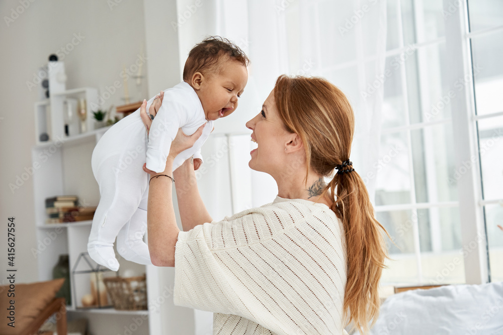 Fototapeta premium Happy young caucasian mother lifting cute little baby daughter. Mum playing with african american infant girl at home. Mixed race mommy and small adorable funny child laughing having fun together.