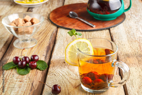 black tea in a glass cup with mint cherries and lemon on a wooden table next to fresh cherries and a teapot on a stand and lemon in a plate.