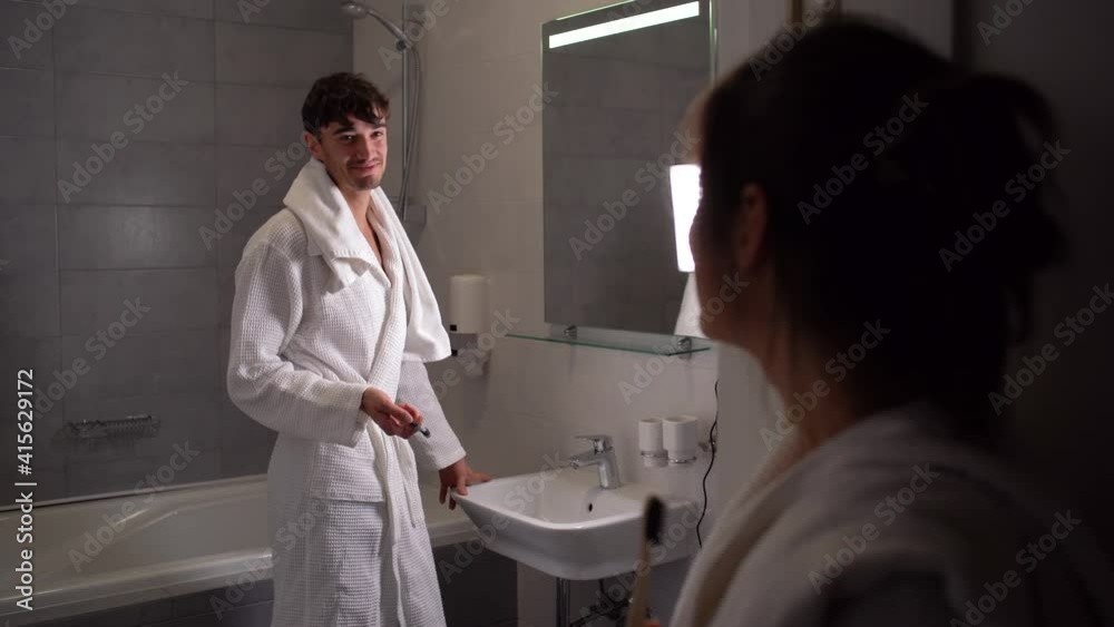 A young European in the bathroom is in a robe and brushes his teeth, his girlfriend watches as the guy takes care of the tooth enamel. A young European in the bathroom is in a robe and brushes his teeth, his girlfriend watches as the guy takes care of the tooth enamel.