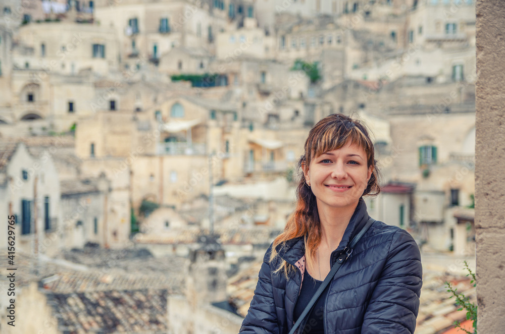 Young beautiful woman traveler with black jacket looking at camera, smile and posing in Sassi di Matera city historical centre Sasso Caveoso with old stone buildings and rock cave houses background