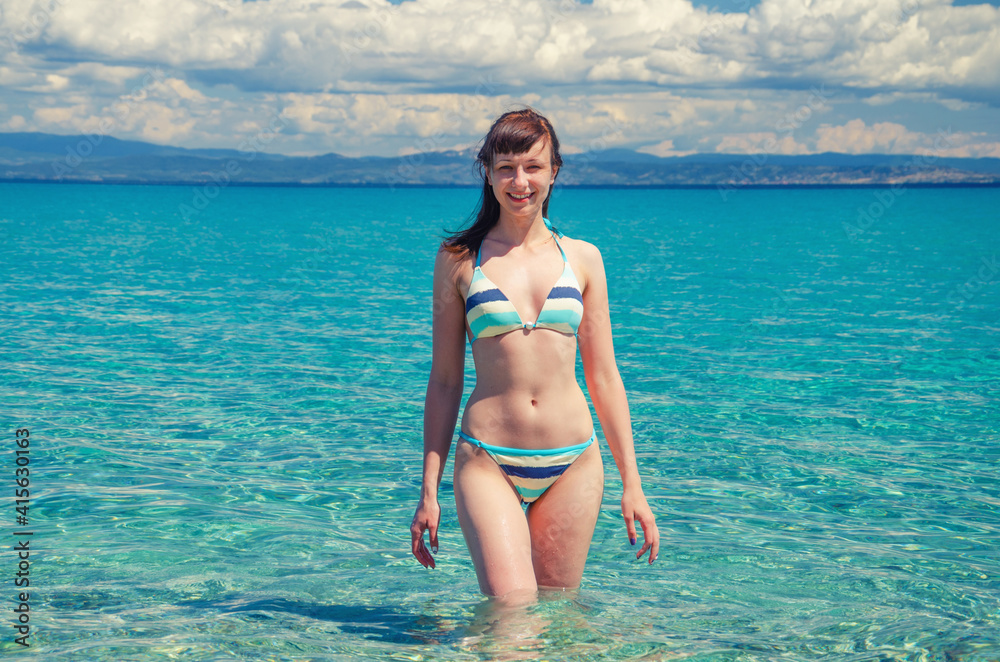 Young beautiful girl in bikini stay in turquoise water of Toroneos kolpos gulf, looking at camera and smile, tourist woman on paradise vacation in Halkidiki of Kassandra Greece in sunny summer day