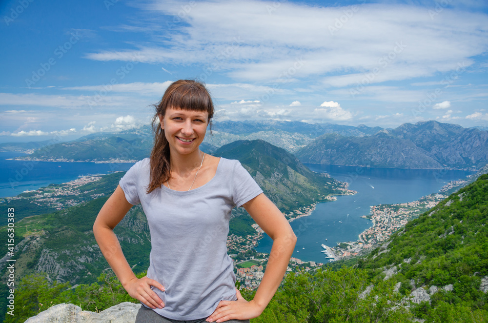 Young beautiful girl traveler with grey t-shirt posing and smile on top of mountain, aerial view of Boka Kotor bay, Old Town Kotor and Dinaric Alps mountains range, woman on vacation in Montenegro