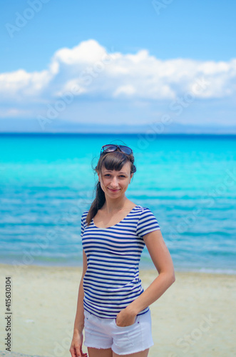 Young beautiful girl with striped shirt and white shorts posing and smile on sandy beach, turquoise water of Toroneos gulf in Halkidiki Kassandra, sunny summer day, vacation in Greece, vertical view