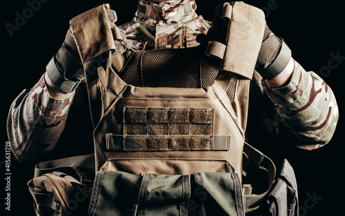 Fototapeta Photo of soldier in camouflaged uniform and tactical gloves holding military armored vest on black background