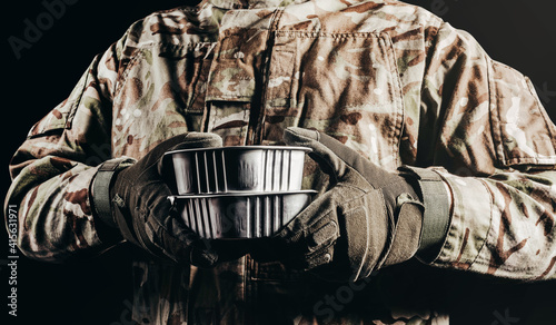 Photo of soldier in camouflaged uniform and tactical gloves holding canned food field ration on black background.