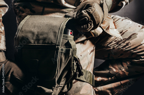 Photo of soldier in camouflaged uniform and tactical gloves using leg bag on black background.