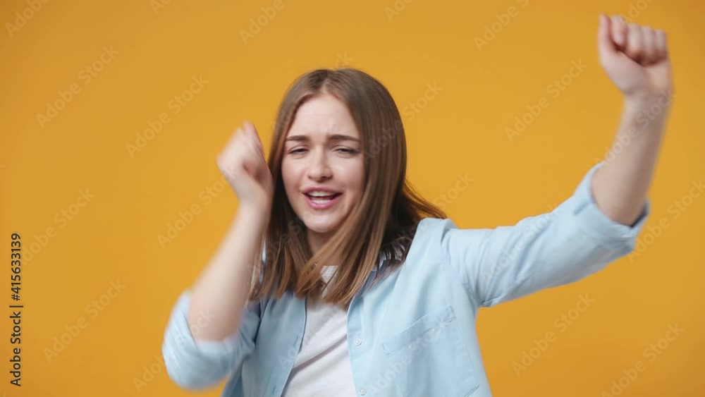 Cheerful funny excited blonde young woman 20s years old in casual blue shirt posing isolated on yellow color background in studio. People lifestyle concept. Dancing clenching fists waving rising hands Cheerful funny excited blonde young woman 20s years old in casual blue shirt posing isolated on yellow color background in studio. People lifestyle concept. Dancing clenching fists waving rising hands
