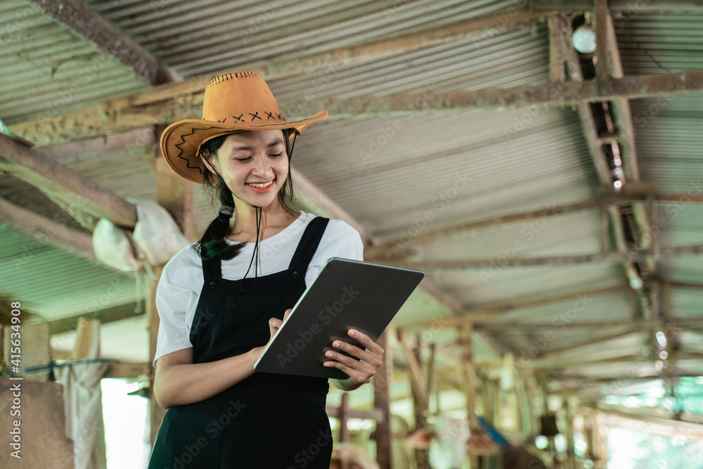 close-up of a smiling woman wearing a cowboy hat while touching a screen of a digital tablet in the background of a cow farm stable