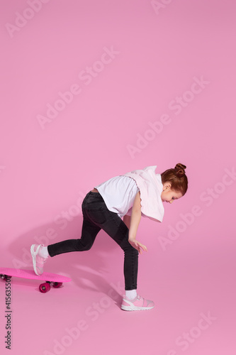 Stylish little child girl is learning to skateboard for the first time over pink background. © producer
