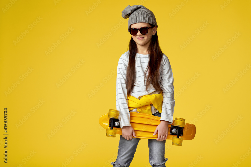 unhappy little child girl in hat and sunglasses posing with yellow skateboard on yellow studio background. unhappy little child girl in hat and sunglasses posing with yellow skateboard on yellow studio background.