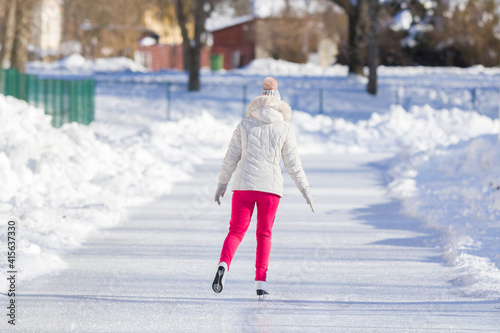 Young adult alone woman in white jacket and pink pants skating on ice track in sunny winter day. Enjoying sport in cold weather. Outdoor activities. Back view. Top down view.