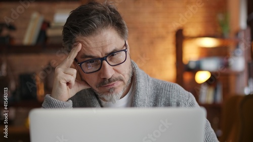 Older man working online with laptop computer at home sitting at desk. Home office, browsing internet, study room. Man thinking solving problem.