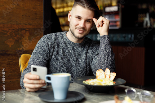 Beautiful smiling male in cafe drink coffee and eating salad. Young man in casual clothes with smartphone in hands.