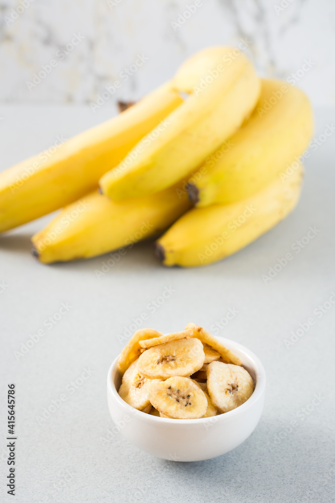 Baked banana chips in a white bowl and a bunch of bananas on the table. Fast food. Close-up. Vertical view Baked banana chips in a white bowl and a bunch of bananas on the table. Fast food. Close-up. Vertical view