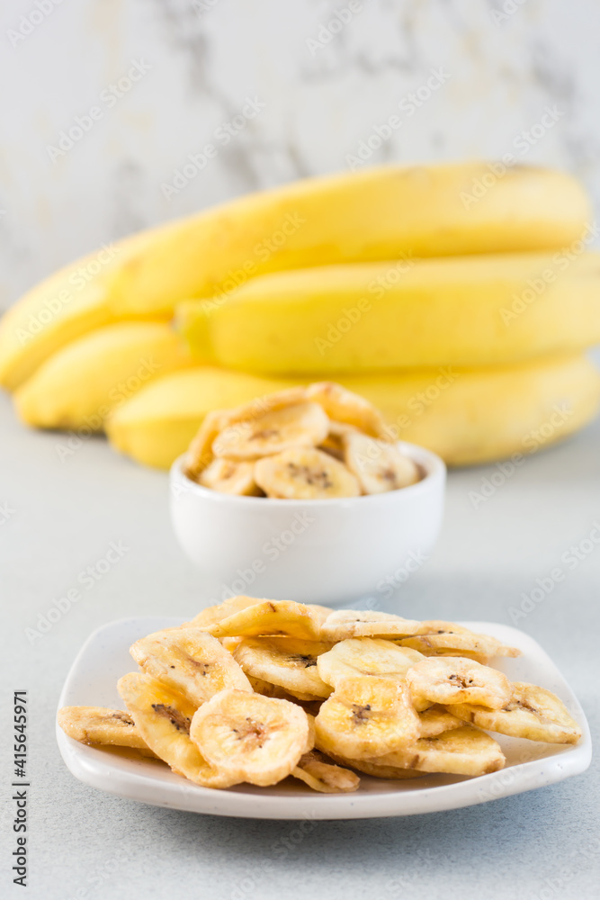 Baked banana chips in a white bowl and saucer and a bunch of bananas on the table. Fast food. Vertical view. Close-up