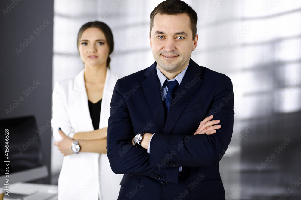 Portrait of a friendly middle aged businessman in a dark blue suit, standing with crossed arms together with a colleague in a modern office. Concept of success in a business Portrait of a friendly middle aged businessman in a dark blue suit, standing with crossed arms together with a colleague in a modern office. Concept of success in a business