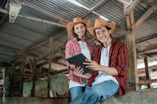 two smiling farmers in casual clothes sit by a wooden fence while using a digital tablet in the background of the cattle farm © Odua Images