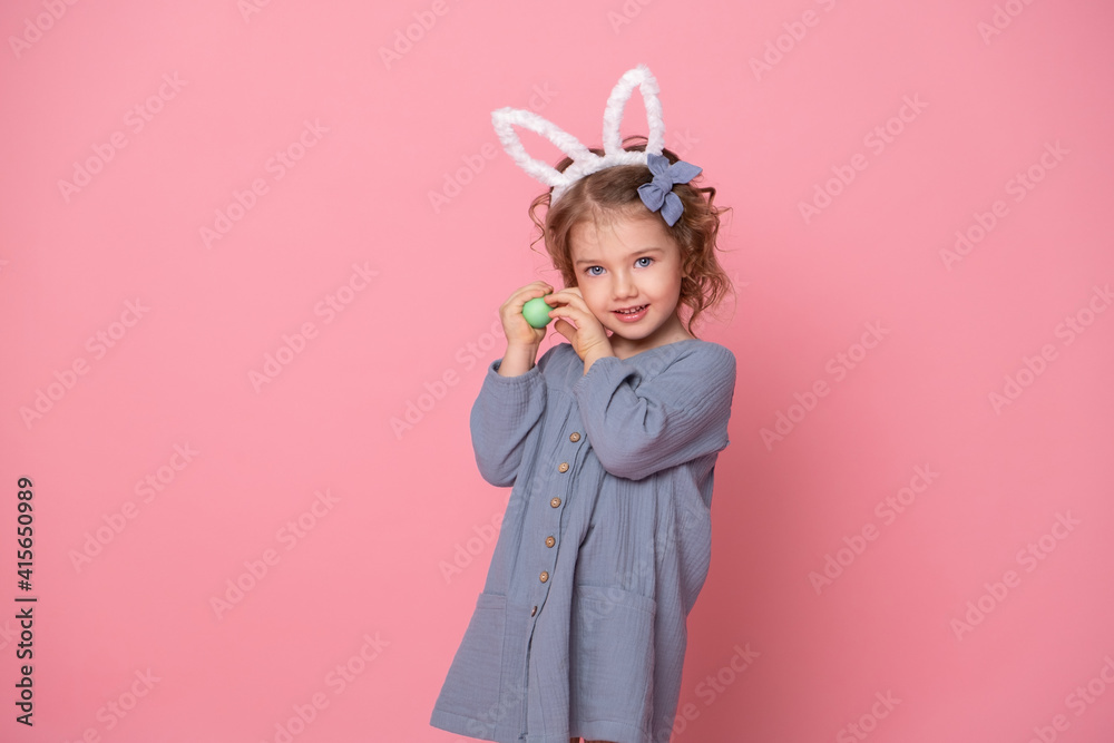 Funny cute child girl in Easter bunny ears and blu dress holding colorful eggs on pink background Funny cute child girl in Easter bunny ears and blu dress holding colorful eggs on pink background