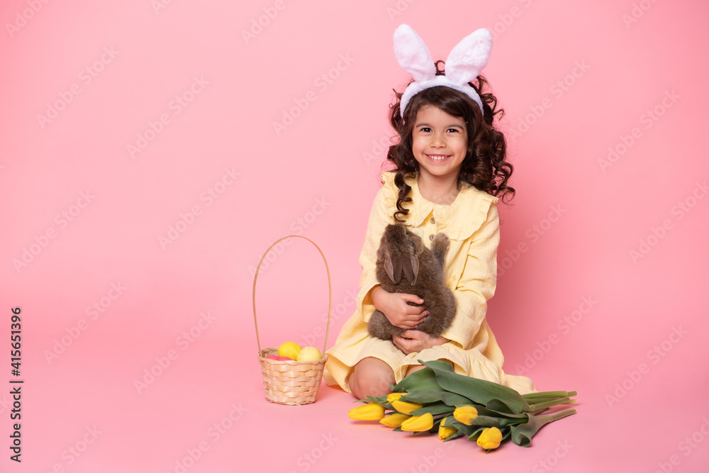 child girl in bunny ears holding rabbit, tulips, basket with colorful eggs on pink background.