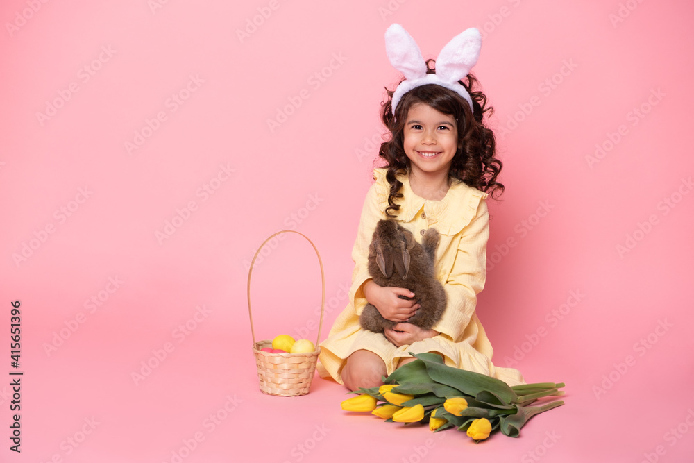 child girl in bunny ears holding rabbit, tulips, basket with colorful eggs on pink background. child girl in bunny ears holding rabbit, tulips, basket with colorful eggs on pink background.