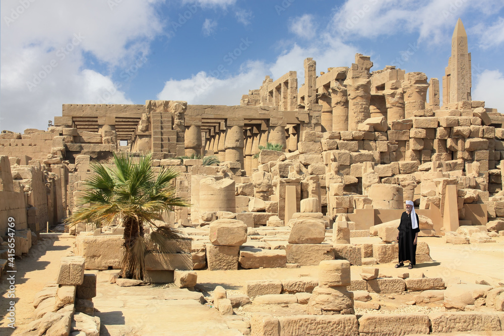An elderly Egyptian stands in the middle of the historical temple complex of Karnak. He wears a black caftan and a turban. On the left is a palm tree.