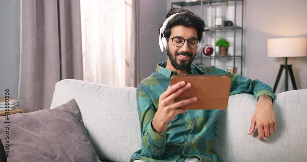 Portrait of Hindu young man watching movie or videos on tablet device in headphones while resting at home sitting in living room. Handsome bearded guy browsing online on gadget. Leisure concept Portrait of Hindu young man watching movie or videos on tablet device in headphones while resting at home sitting in living room. Handsome bearded guy browsing online on gadget. Leisure concept