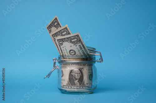 A glass jar full of dollars between one dollar bills. An easy way to save real money. Blue background. Bank image and photo.