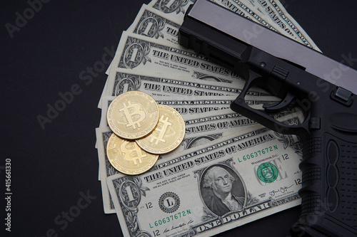 Pile of US dollars cash and a black pistol. Next to it are several gold bitcoin electronic cryptocurrency coins. Seen from above.