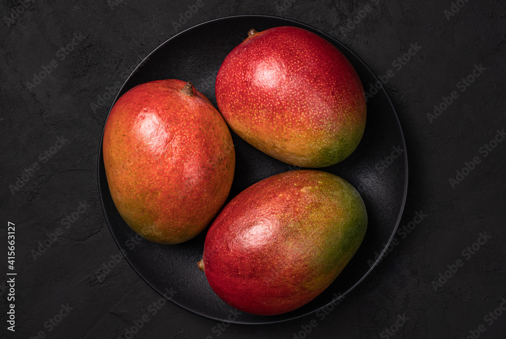 Three mango fruits on the black plate.Top view.Dark food phhotography. Three mango fruits on the black plate.Top view.Dark food phhotography.