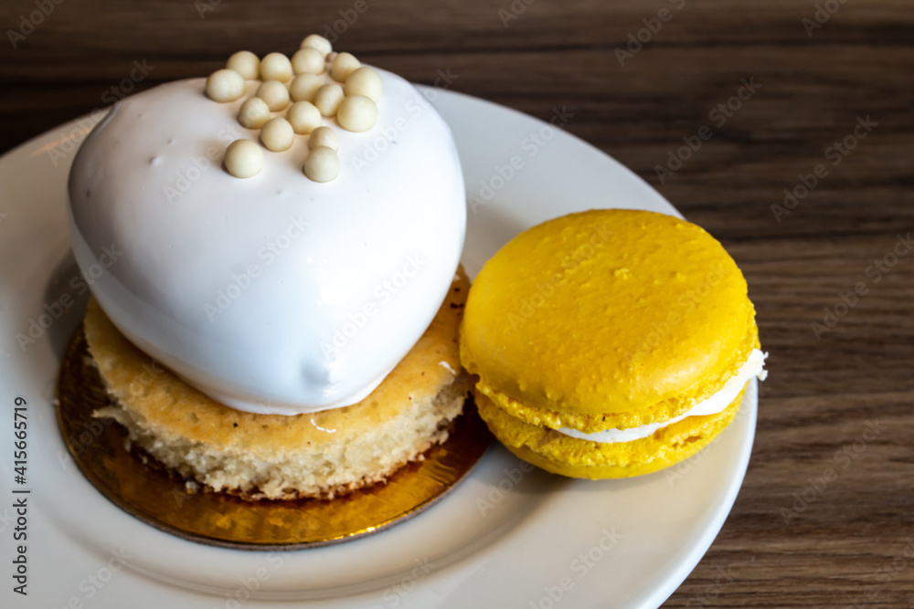 Heart-shaped vanilla passionfruit cake on a gold foil circle, February 2021. Mango and Passionfruit cream-filled French macaron on the side of a white ceramic plate, hardwood table background.