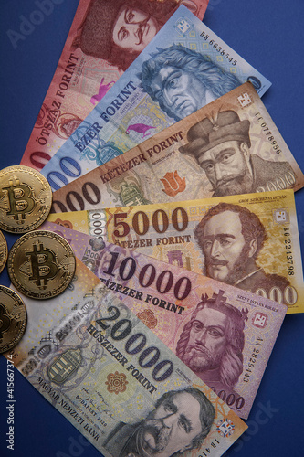 Several Hungarian forint paper money denominations with a gold bitcoin digital cryptocurrency coin on a blue background. Bank image and photo.