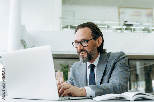 Smiling mature male employee is chatting with colleagues uses a laptop. Successful businessman working on new project, searching ideas, browsing internet
