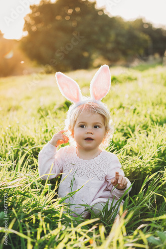 Happy and cute baby girl wears rabbit ears. Sitting in green grass. The girl has blonde curly hair and a pink dress. Easter concept. Spring time. 