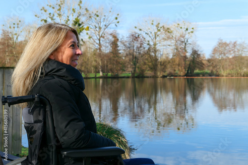 Concept of disabled person. A woman laughing in a wheelchair outside in the nature in front of a lake.  © Bruno
