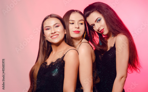 3 girls posing, laughing on a pink background. Girls in brilliant dresses with different colorful makeup. Three brunette girls dance and enjoy life. March 8, women's power, Women's Day