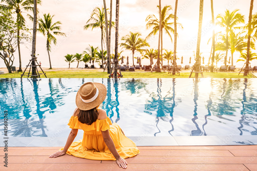 Young woman traveler relaxing and enjoying the sunset by a tropical resort pool while traveling for summer vacation - obrazy, fototapety, plakaty 