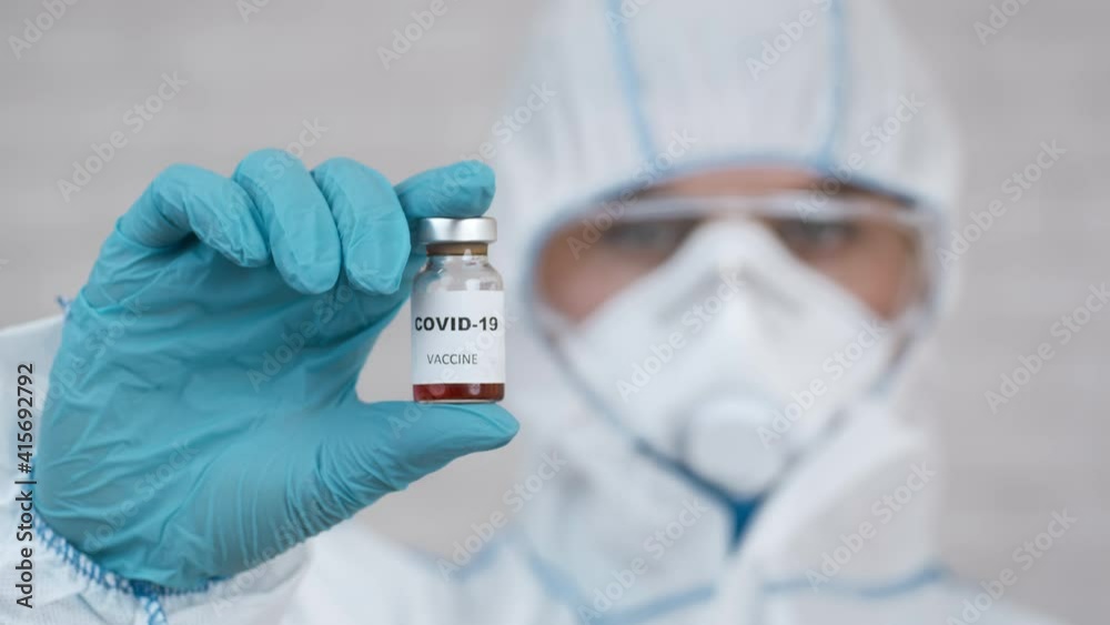Doctor Holding Covid-19 Vaccine Bottle Indoors, Wearing Protective Hazmat Suit Doctor Holding Covid-19 Vaccine Bottle Indoors, Wearing Protective Hazmat Suit