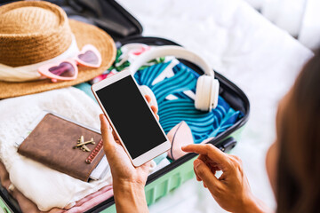 Young woman traveler sitting on the bed using smartphone and packing her suitcase preparing for travel on summer vacation