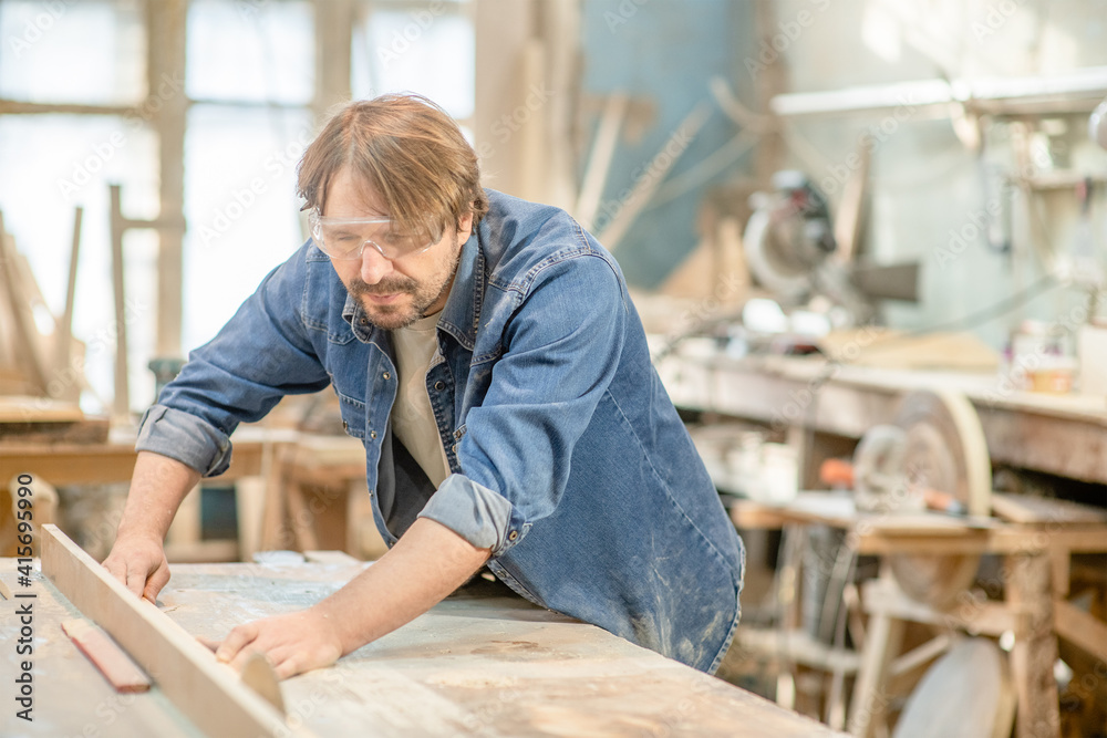 Carpenter wearing safety googles cutting a piece of wood at workshop, using a circular saw