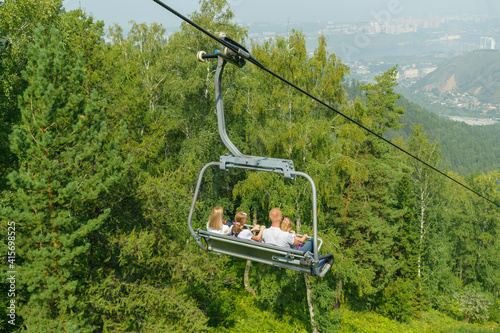 Family rides the chairlift down. View from the back. Background - green forest, city. The concept of summer family vacation in the mountains, vacations.