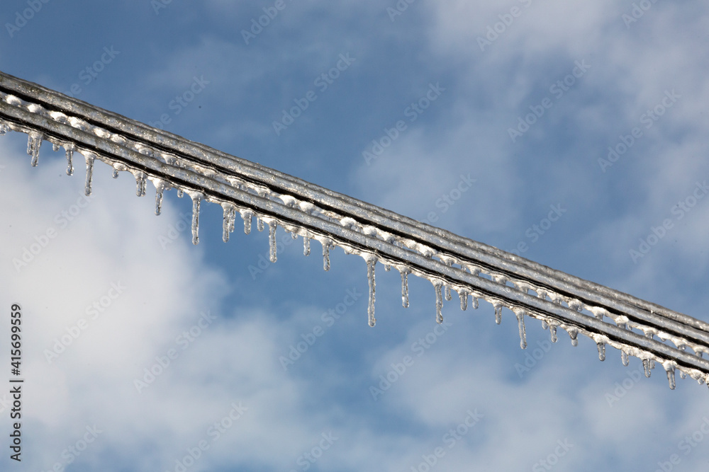Two parallel electrical wires covered with ice and icicles against a blue sky with white wispy clouds
Two parallel electrical wires covered with ice and icicles against a blue sky with white wispy clouds