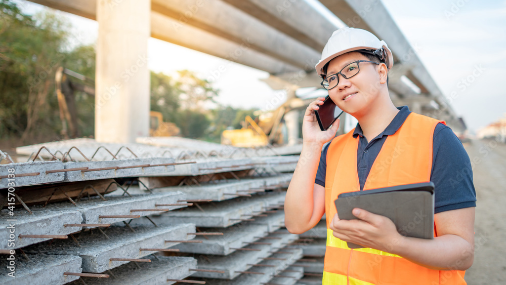 Smart Asian worker man or male civil engineer with protective safety helmet and reflective vest using digital tablet for project planning and checking material at construction site.