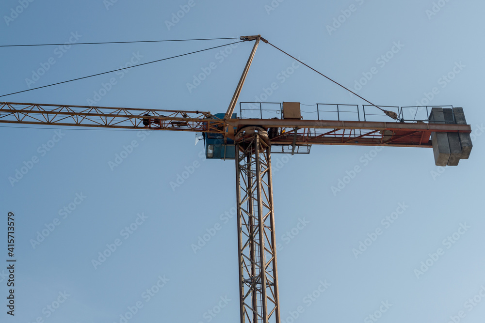 Fragment of upper part of yellow tower crane with reinforced frame, mesh floor, blue cab,  boom extending to left and counterweight of concrete slabs on right, between which cables stretched.