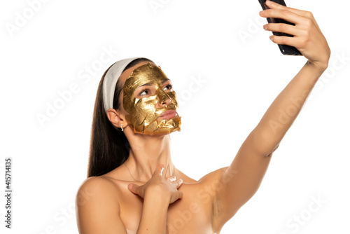 young woman takes selfies with golden mask on her face on white background
