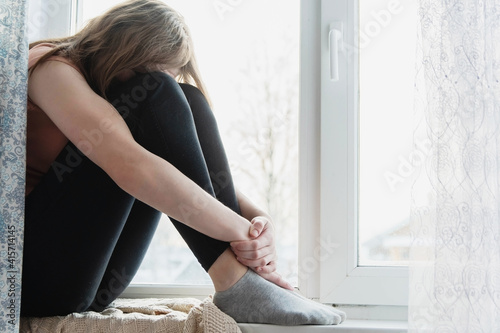 The teenage girl sits on the window sill bending her legs and burying her face in her knees. Concept of depression, loneliness in teenagers.
