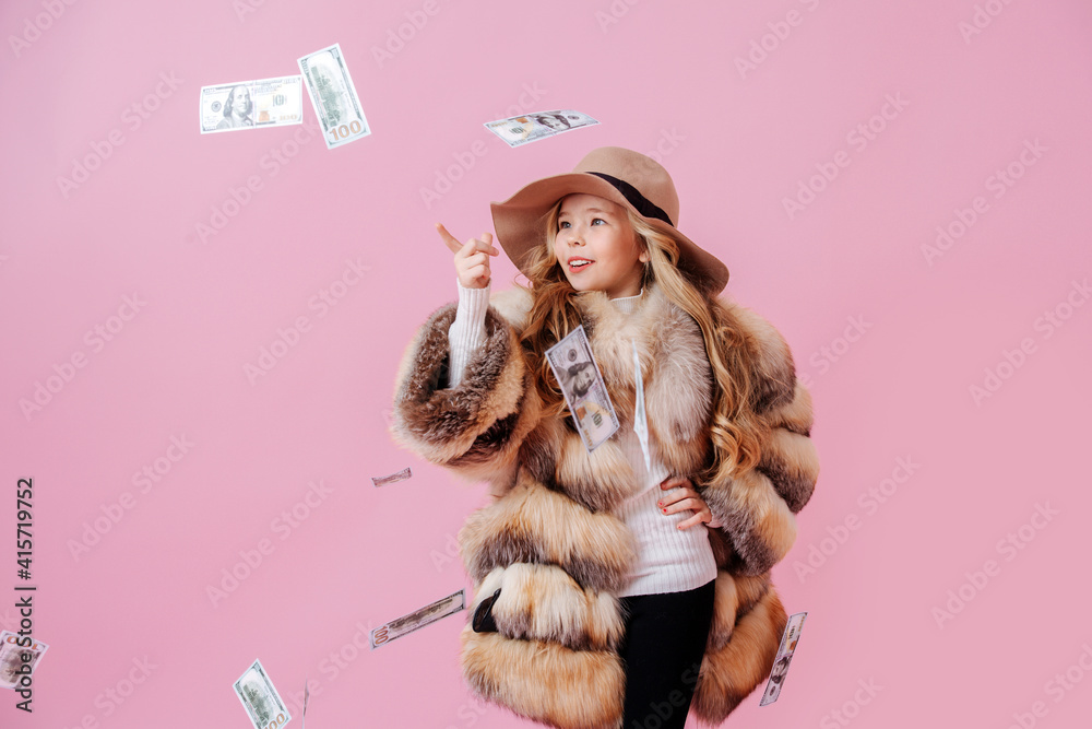 Pretty preteen girl in oversized luxurious fur coat and pole hat pointing finger at money raining at her. Over dark pink background.