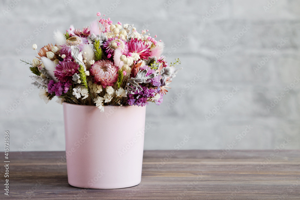 A delicate bouquet of dried flowers in a pink vase. Wooden surface, brick wall. Close-up, copy space. The concept of a gentle greeting card in pastel colors