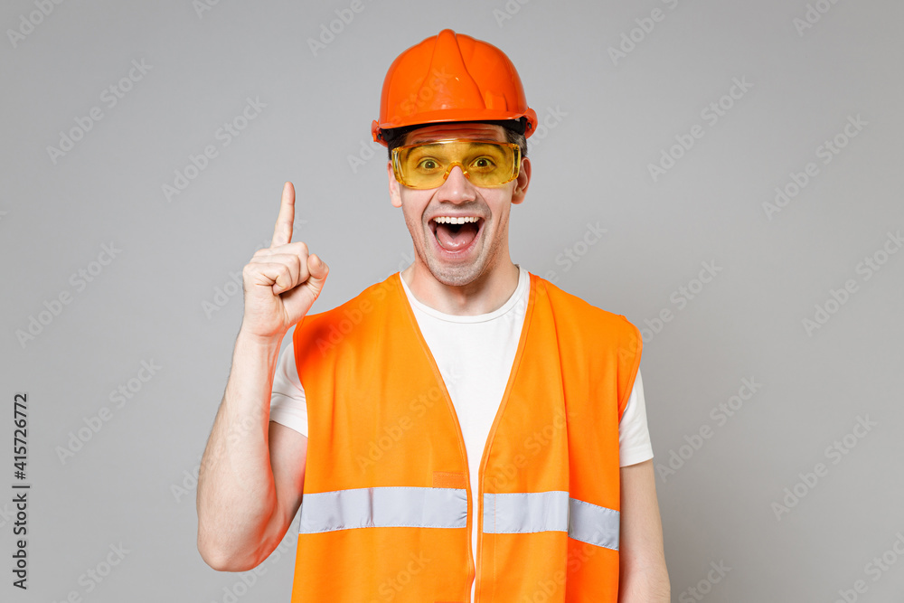 Young employee man in orange vest protective hardhat holding index finger up, great new idea isolated on grey background studio portrait. Instruments for renovation apartment room Repair home concept Young employee man in orange vest protective hardhat holding index finger up, great new idea isolated on grey background studio portrait. Instruments for renovation apartment room Repair home concept